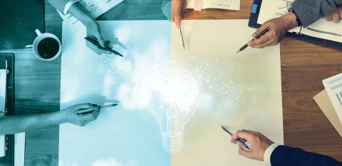 Overhead photo of people's hands pointing to a lightbulb on a meeting table